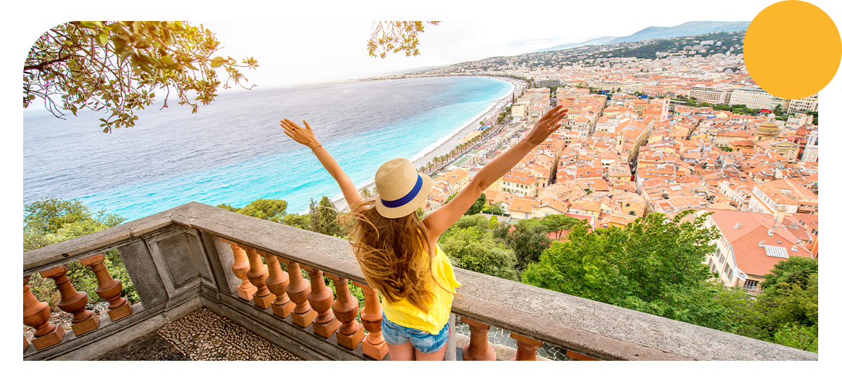 Photo d'une femme les bras écarté, située sur la colline du chateau à Nice, avec la ville en fond